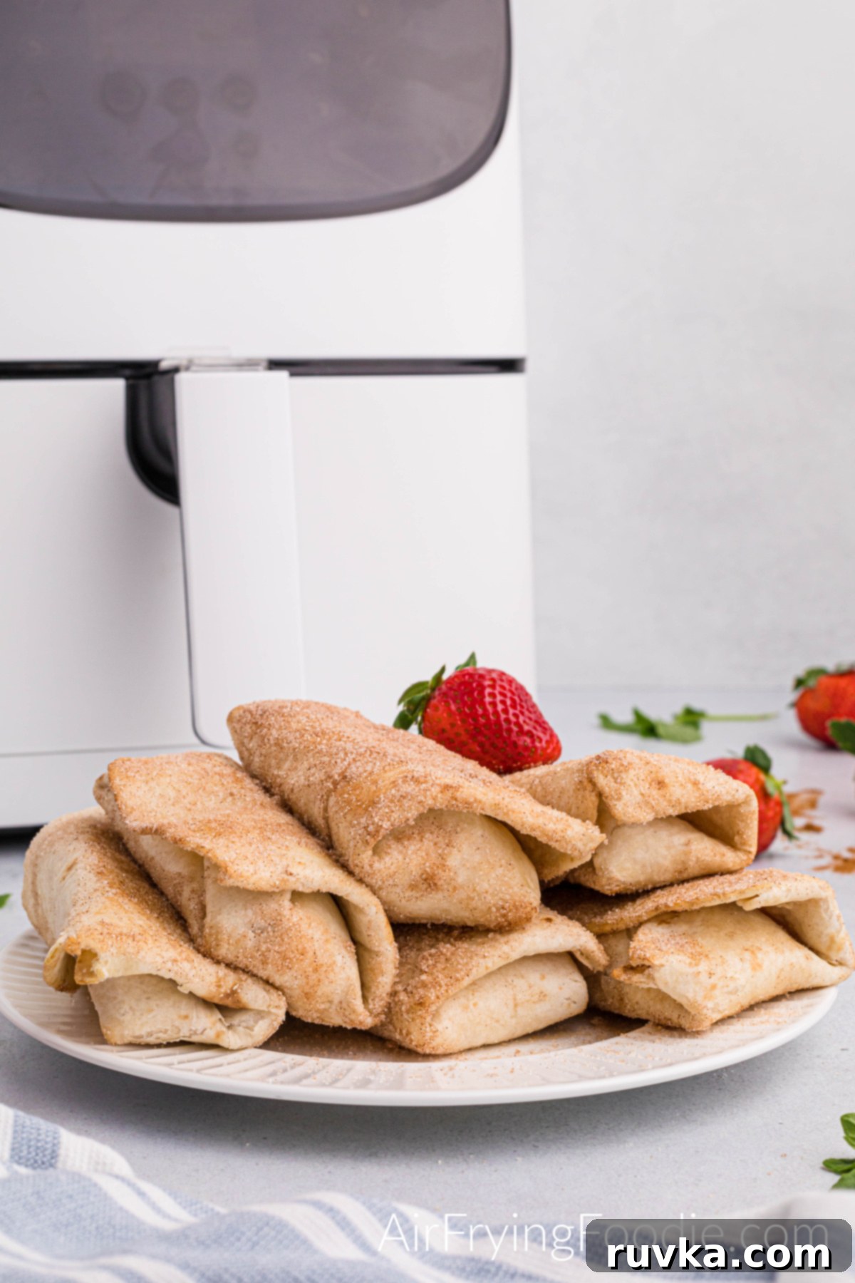 Crispy Air Fryer Strawberry Cheesecake Chimis 15 Round white plate with six strawberry cheesecake chimichangas cooked in the air fryer topped with cinnamon and sugar with a whole strawberry on top of the chimichangas. The base of the air fryer is behind the plate, and whole strawberries are on the table to the right.