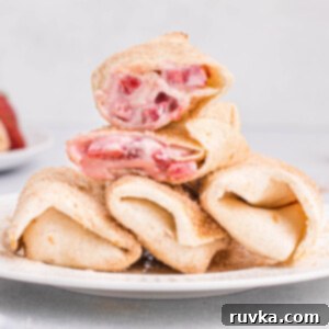 Crispy Air Fryer Strawberry Cheesecake Chimis 16 Air fryer strawberry cheesecake chimichangas served on a round white plate. There are three chimichangas lined on the bottom of the plate. One of the chimichangas is cut in half and stacked on top.
