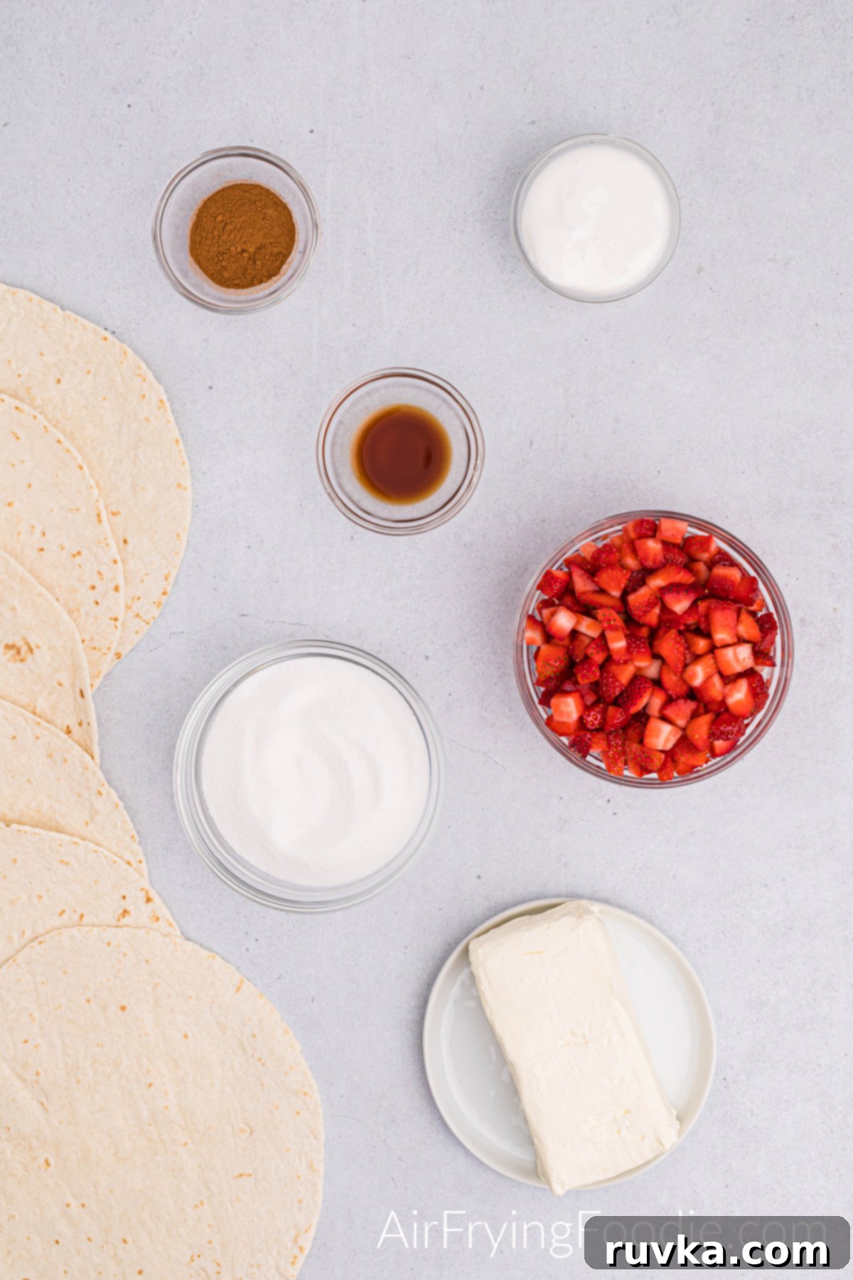 Crispy Air Fryer Strawberry Cheesecake Chimis 3 Ingredients shot with everything you need to make air fryer strawberry cheesecake chimichangas. To the left of the picture is a line of tortillas. The bottom of the image has a small rimmed white plate with a block of cream cheese. The rest of the photograph are bowls, each containing an ingredient; sugar, cut-up strawberries, vanilla extract, cinnamon, and marshmallow cream.