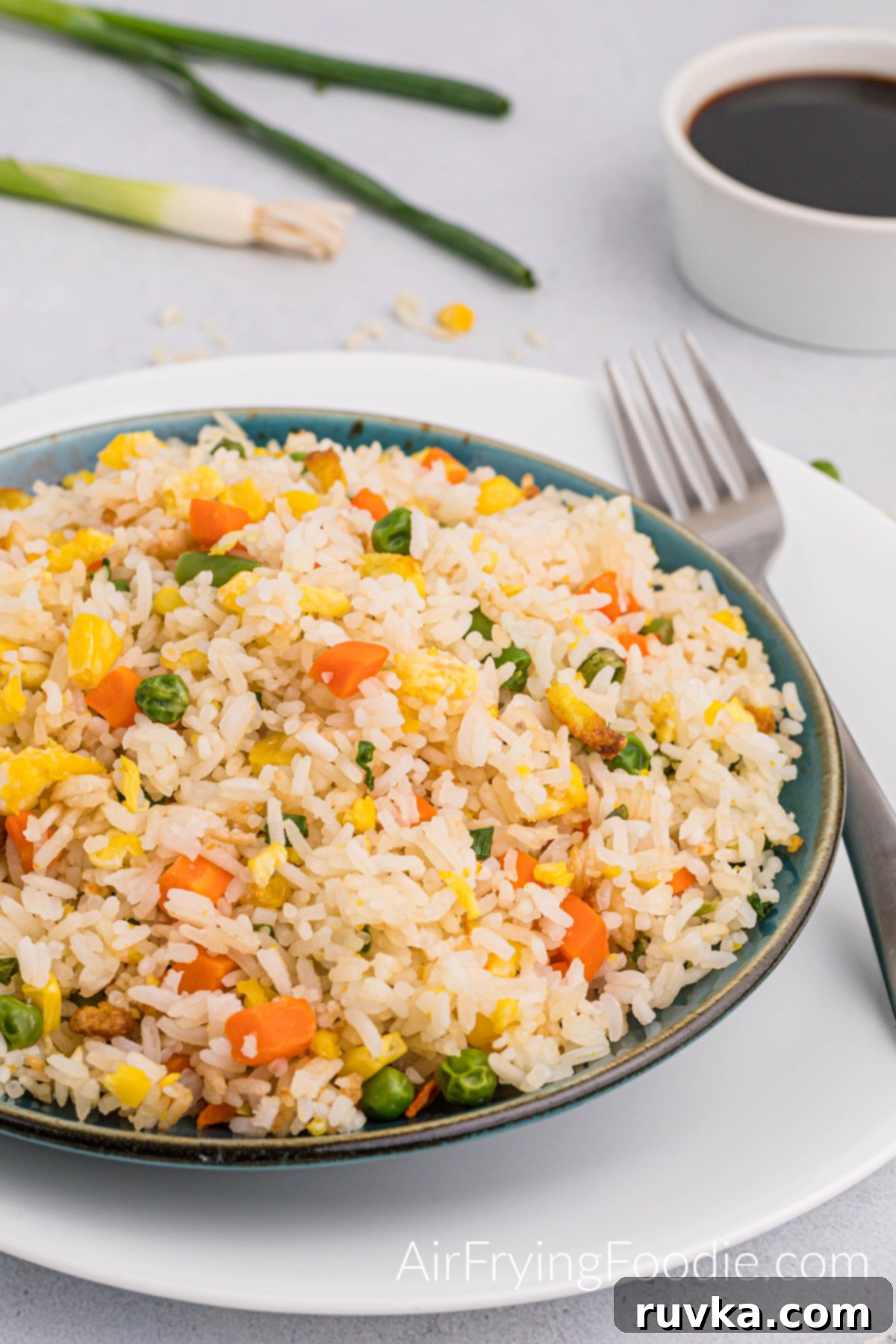 Quick Air Fryer Fried Rice 2 A vibrant bowl of freshly cooked Air Fryer Fried Rice, garnished generously with green onions, served on a round plate. The colorful mix of tender vegetables, scrambled eggs, and fluffy rice is perfectly complemented by a fork and a small bowl of soy sauce in the background.