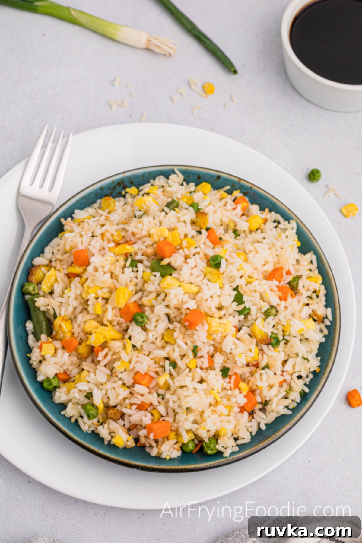 Quick Air Fryer Fried Rice 12 An overhead shot of a vibrant serving of air fryer fried rice on a round plate with a distinctive green rim. A fork is placed beside the plate, and a small white bowl holding soy sauce and green onions completes the fresh and inviting presentation.