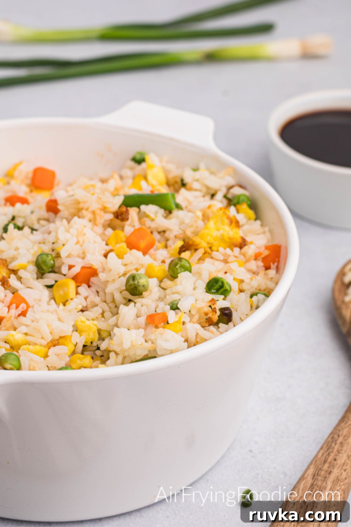 Quick Air Fryer Fried Rice 13 A large white serving dish brimming with air fryer fried rice, beautifully garnished with fresh green onions. Adjacent to the bowl are a wooden serving spoon and a small white bowl of soy sauce, with whole green onions artfully placed on the table in the background.