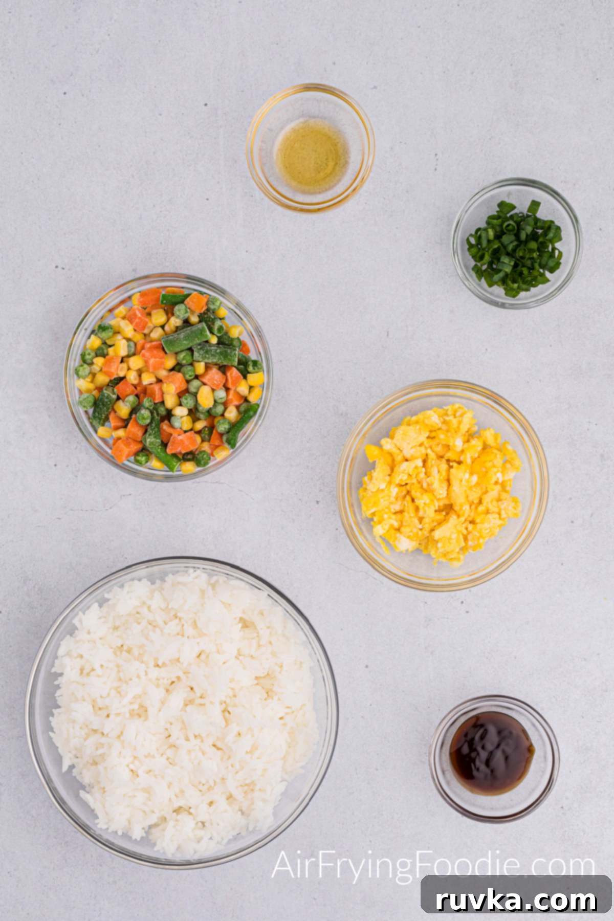 Quick Air Fryer Fried Rice 3 An overhead shot displays the fresh ingredients required for air fryer fried rice: separate glass bowls hold sesame oil, green onions, and oyster sauce. Larger bowls feature vibrant mixed vegetables, fluffy scrambled eggs, and a generous portion of white rice.