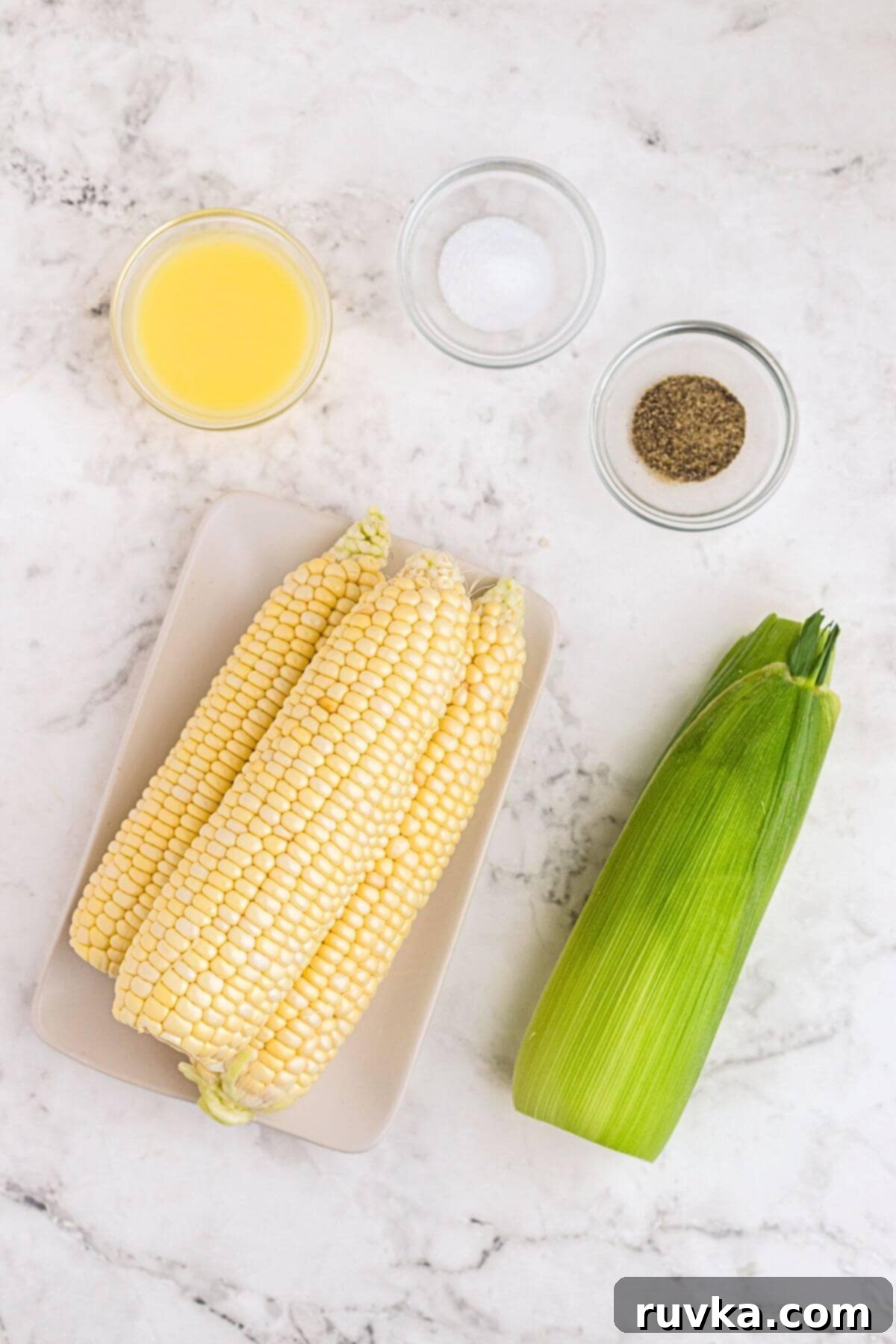 Crispy Air Fryer Corn 3 Uncooked corn on a white marble table with seasonings and melted butter, ready to be prepared.