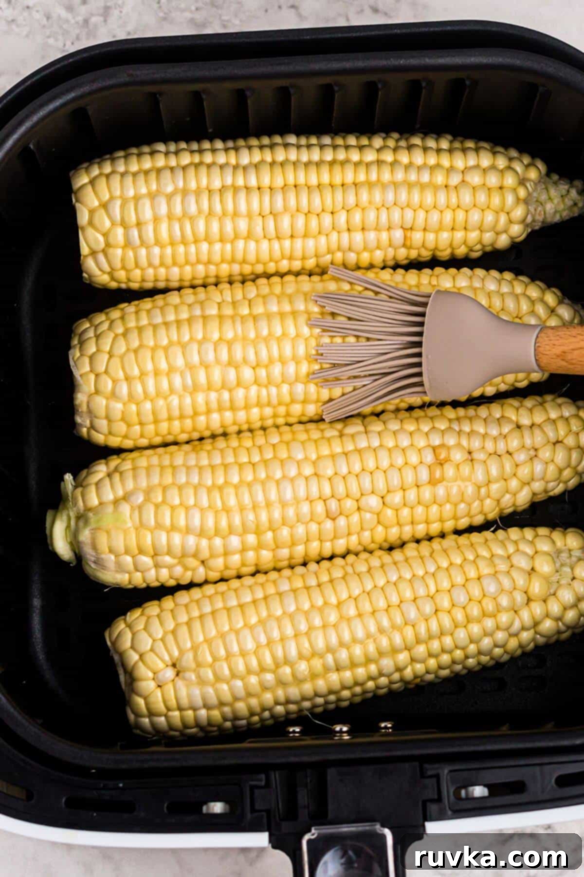 Crispy Air Fryer Corn 4 Ears of corn in the air fryer basket, being brushed with melted butter before cooking to ensure even coating.