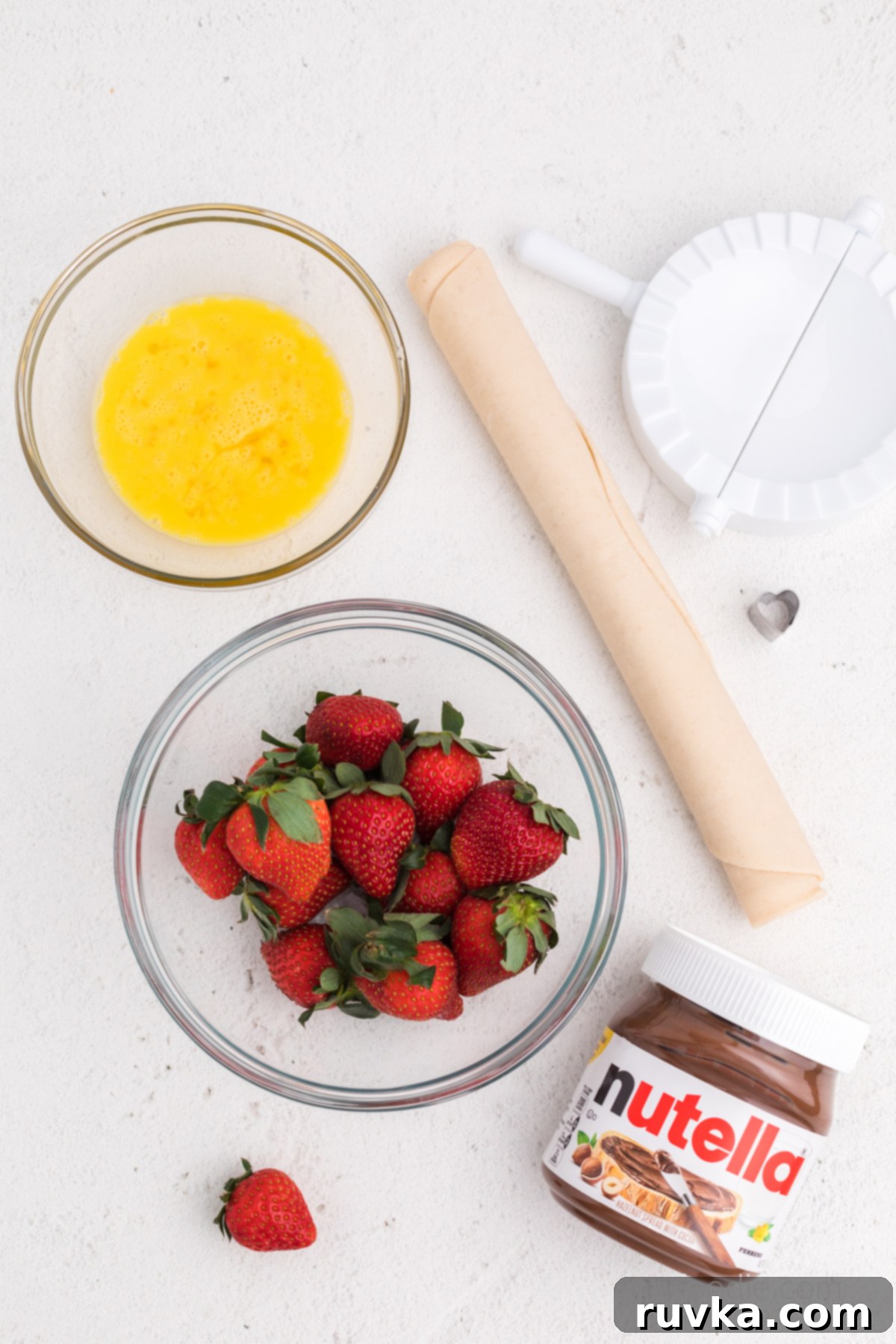 Easy Air Fryer Strawberry Nutella Hand Pies 3 Bowls of fresh sliced strawberries, creamy Nutella, a sheet of premade pie crust, and a whisked egg, showcasing the simple ingredients.