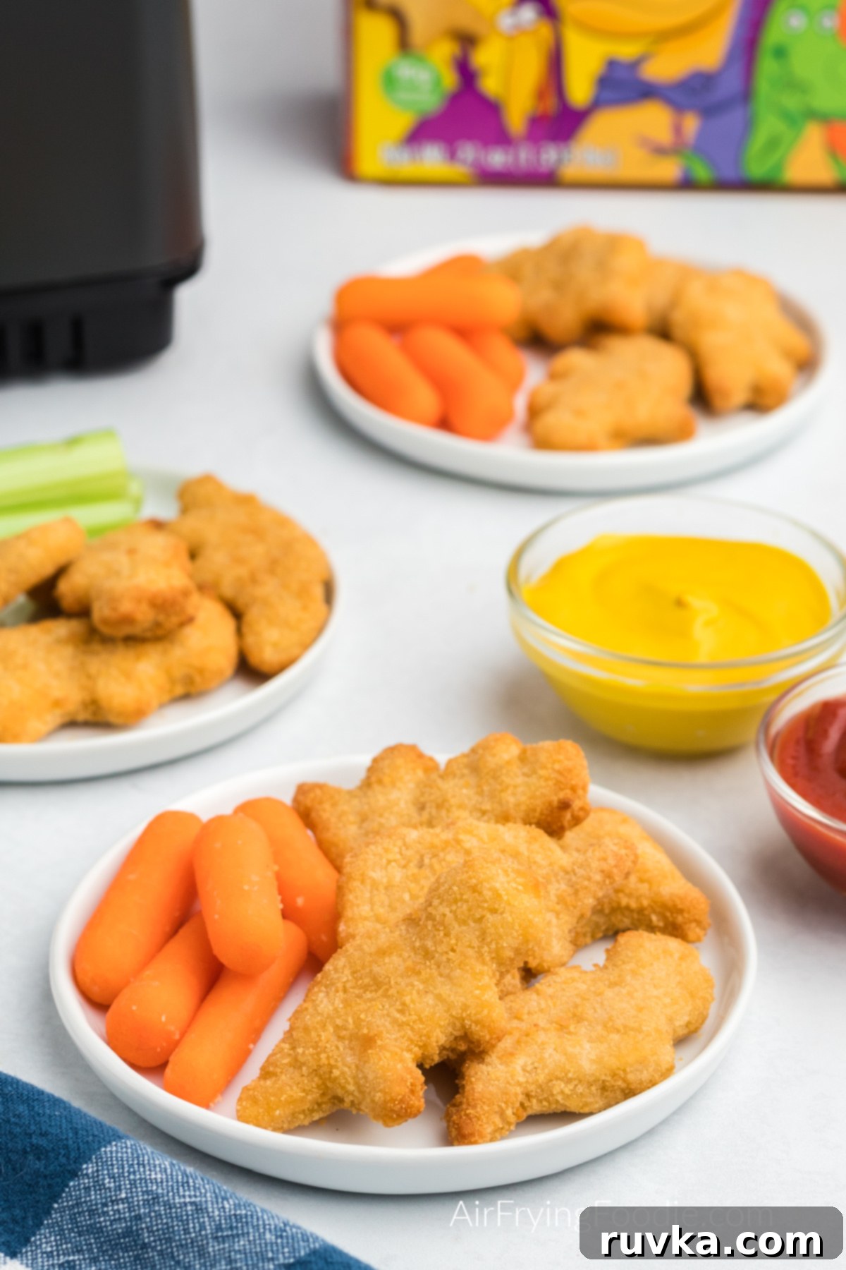 Crispy Air Fryer Dino Nuggets 2 Dino nuggets made in the air fryer, served on plates with carrots and sides of ketchup and mustard.