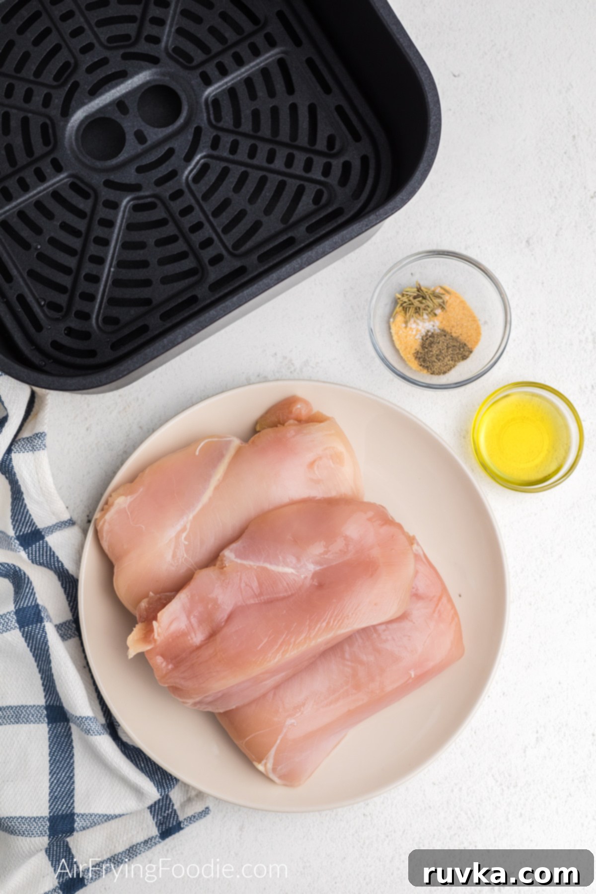 Juicy Air Fryer Grilled Chicken Breast 3 Boneless skinless chicken breasts on a white plate with a small bowl of olive oil and a bowl of seasonings.