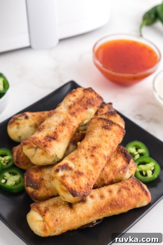 Air Fryer Jalapeno Popper Crunch Rolls 2 Golden crispy Air Fryer Jalapeno Popper Egg Rolls on a black plate, garnished with fresh jalapeno slices.