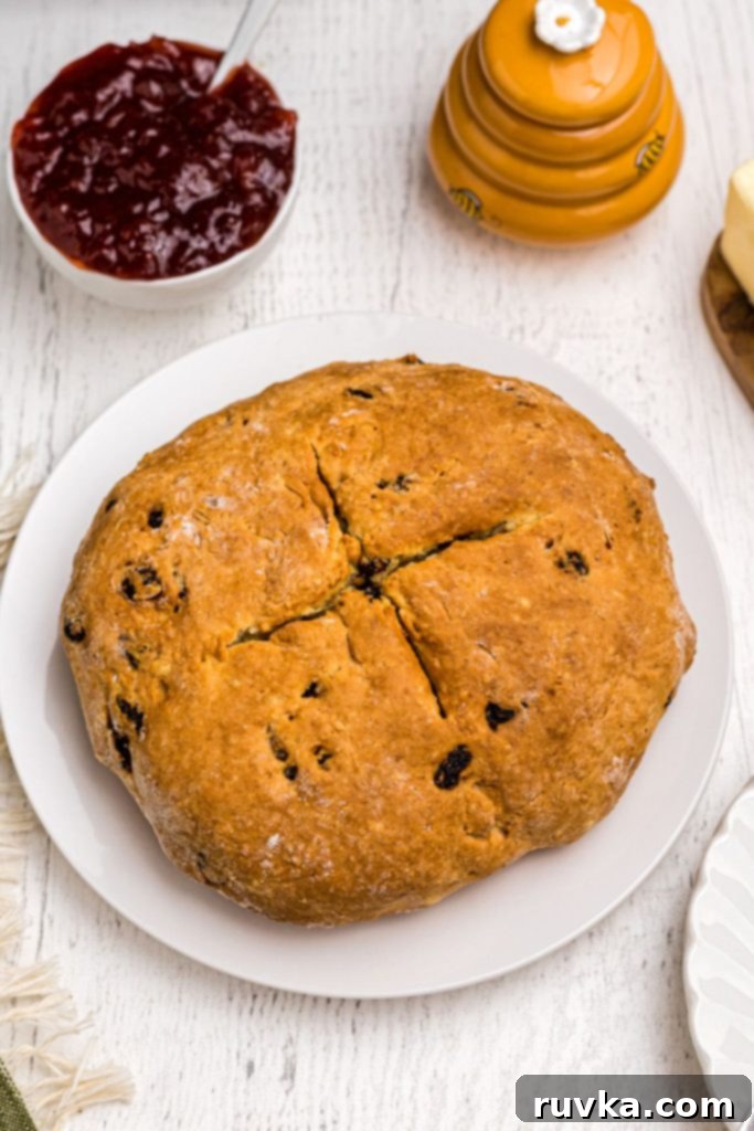 Easy Air Fryer Irish Soda Bread 2 Golden brown round loaf of bread with raisins in it, on a white plate surrounded by butter, honey, and jam