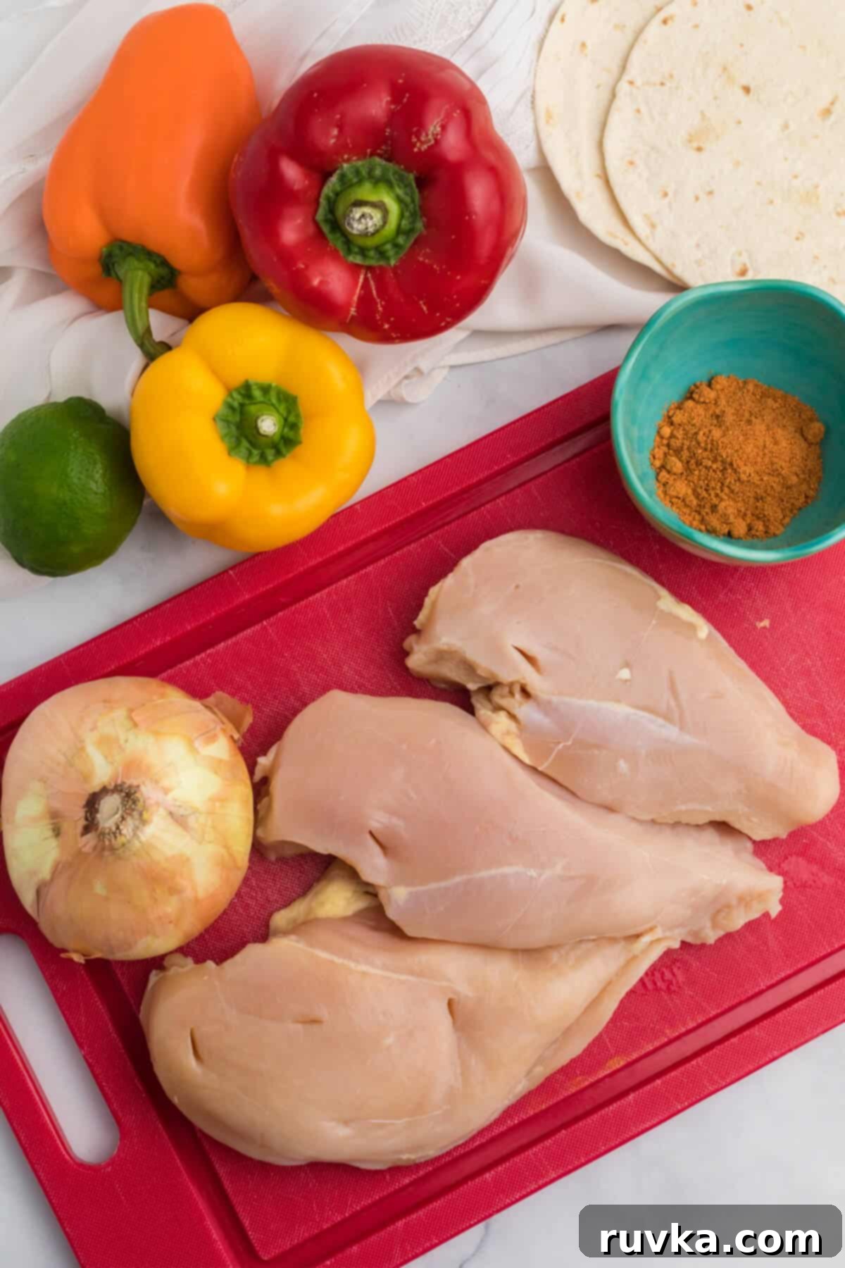 Easy Air Fryer Chicken Fajitas 3 bell peppers, lime, flour tortillas, and a red cutting board with an onion, chicken breasts, and a bowl of taco seasoning.