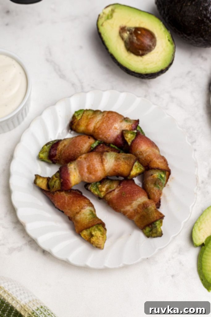 Air Fryer Bacon Wrapped Avocado Perfection 2 Crispy bacon wrapped around avocado wedge on a white plate on a marble table.