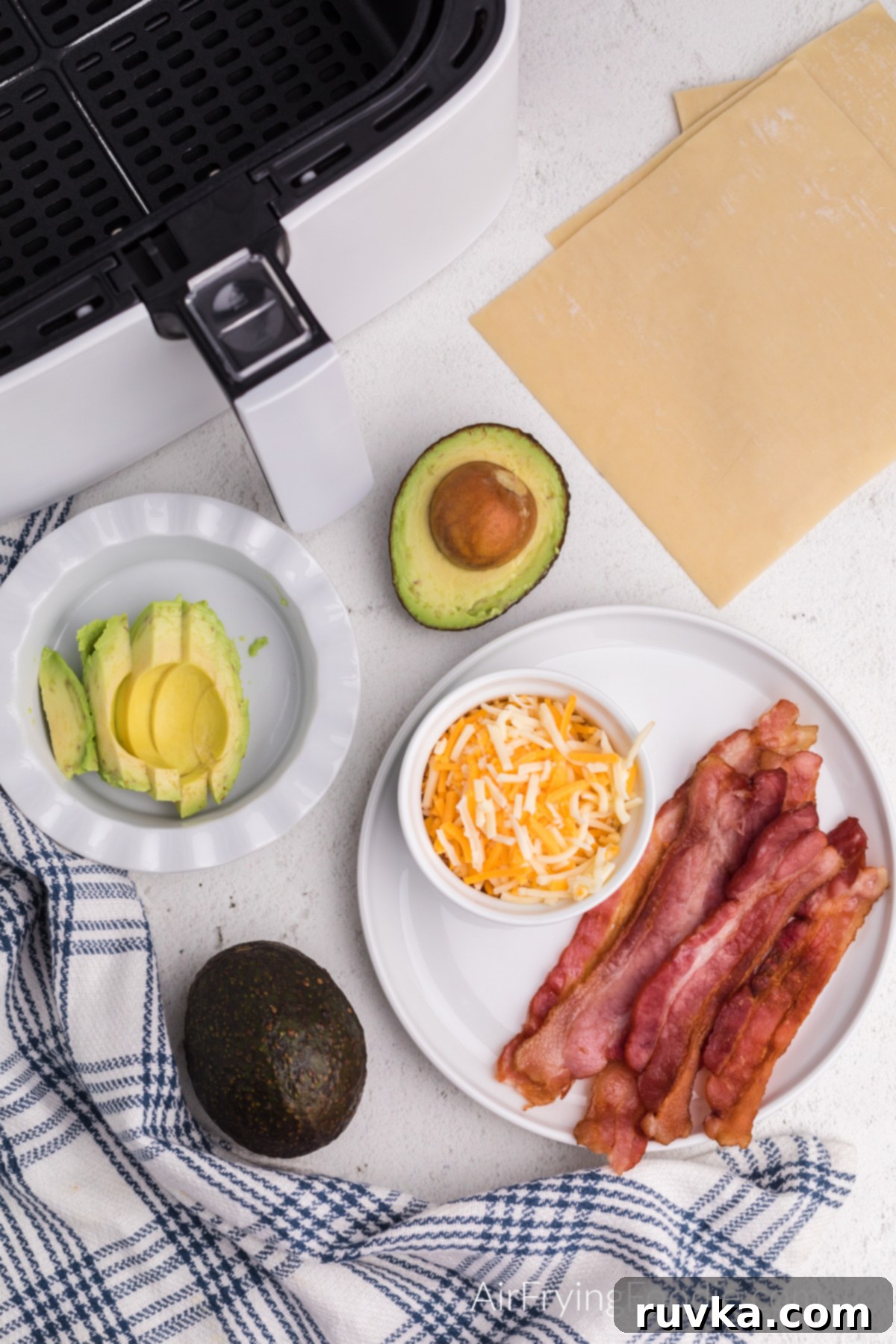 Crispy Air Fryer Avocado Bacon Egg Rolls 3 Ingredients laid out: a plate with cooked bacon, a bowl with shredded cheese, a bowl with sliced avocado, and a stack of egg roll wrappers with whole avocados in the background.