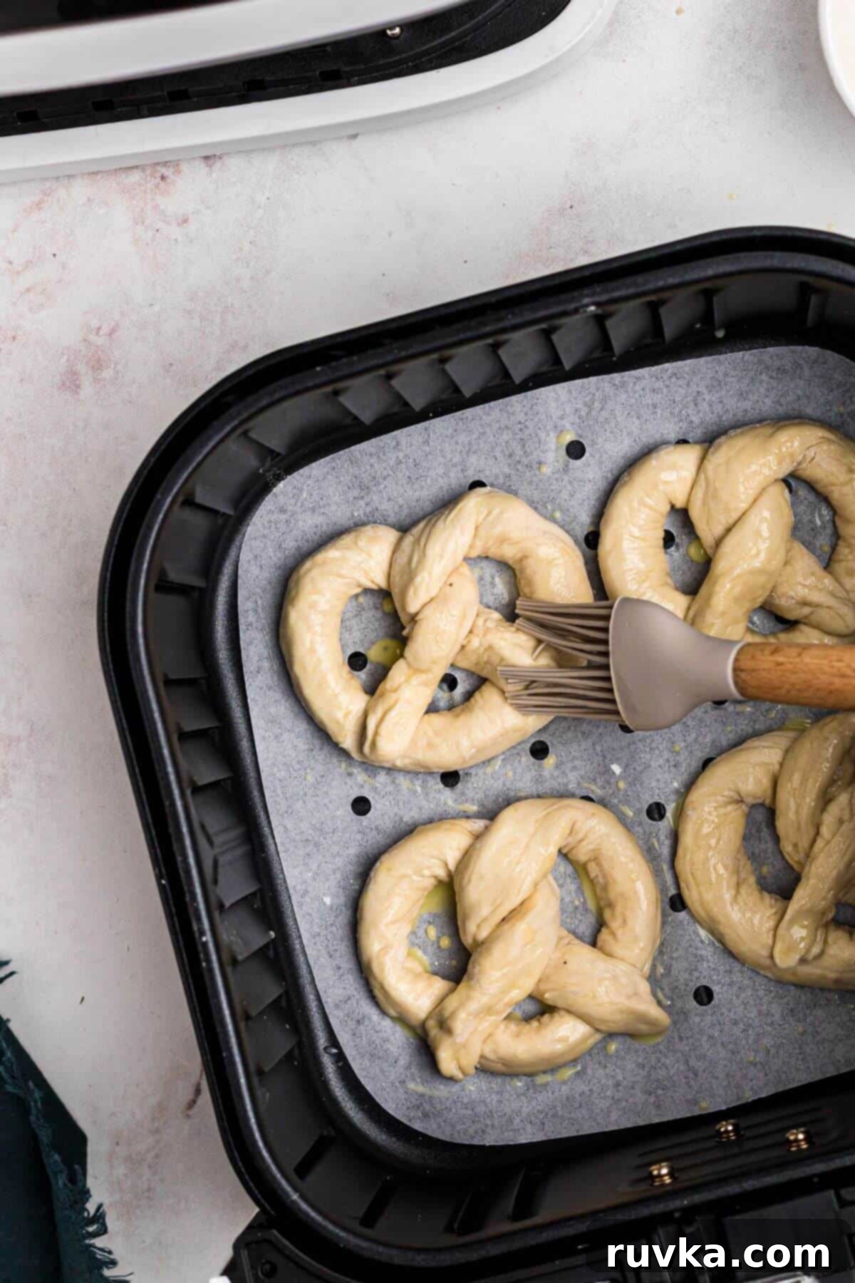 Perfectly Soft Air Fryer Pretzels 8 Uncooked pretzels in the air fryer basket being brushed with egg wash.