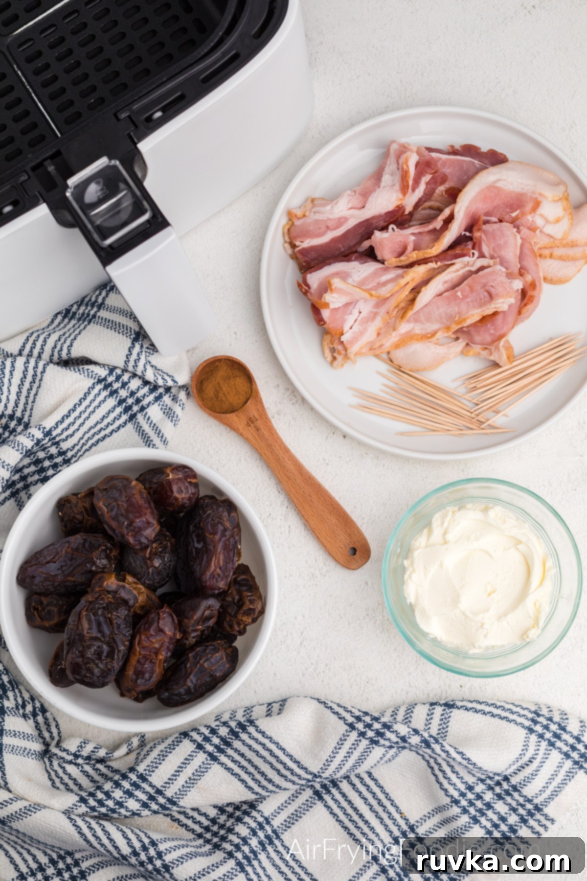 Crispy Air Fryer Bacon Wrapped Dates 3 Table with air fryer basket, bowl of dates, cream cheese, plate with bacon and toothpicks, and a teaspoon of cinnamon.