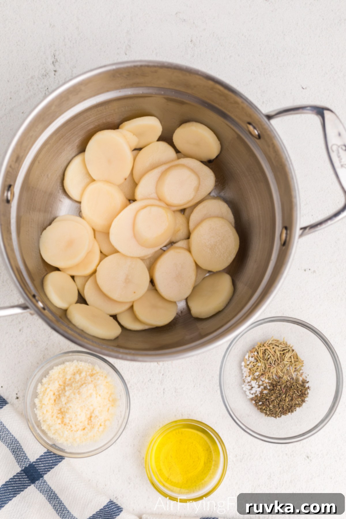 Golden Crispy Air Fryer Potatoes 3 Sliced potatoes in a colander, grated parmesan cheese in a bowl, a small bowl of mixed dried herbs (rosemary, thyme, sea salt), and olive oil in a small bowl, all ingredients ready for preparation.