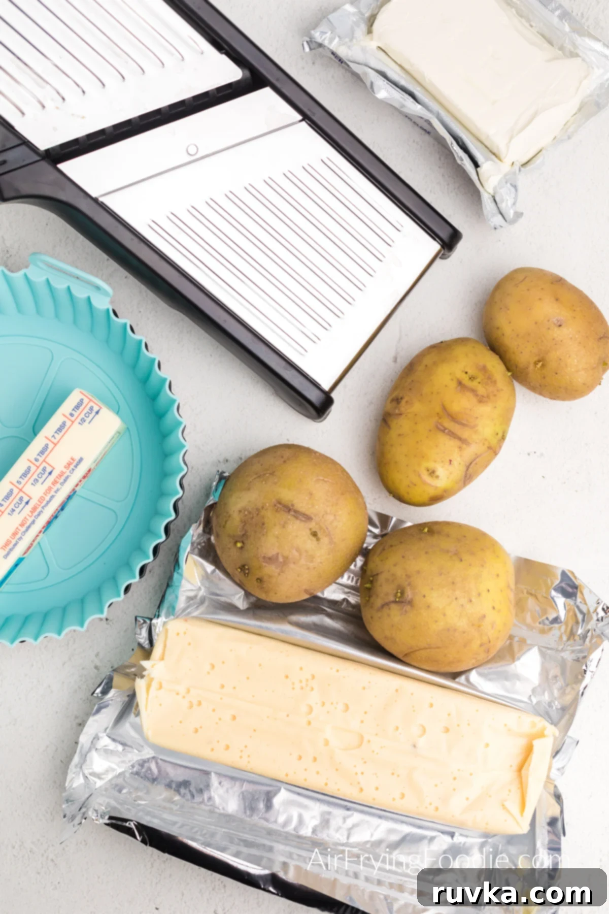 Easy Cheesy Air Fryer Scalloped Potatoes 3 Mandolin slicer, silicone baking dish, cream cheese, butter, queso block cheese, and whole potatoes, neatly arranged on a kitchen counter.