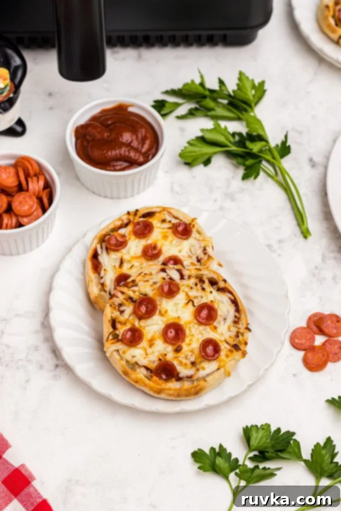 Easy Air Fryer English Muffin Pizzas 9 Two golden crispy English Muffin Pizzas on a small white plate, adorned with a rich pizza sauce and savory pepperonis, all set against a chic marble table background.