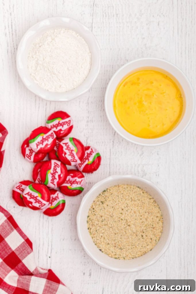 Crispy Air Fried Babybel 3 Babybell cheese wheels, breadcrumbs, whisked eggs, and flour on a white wooden table.