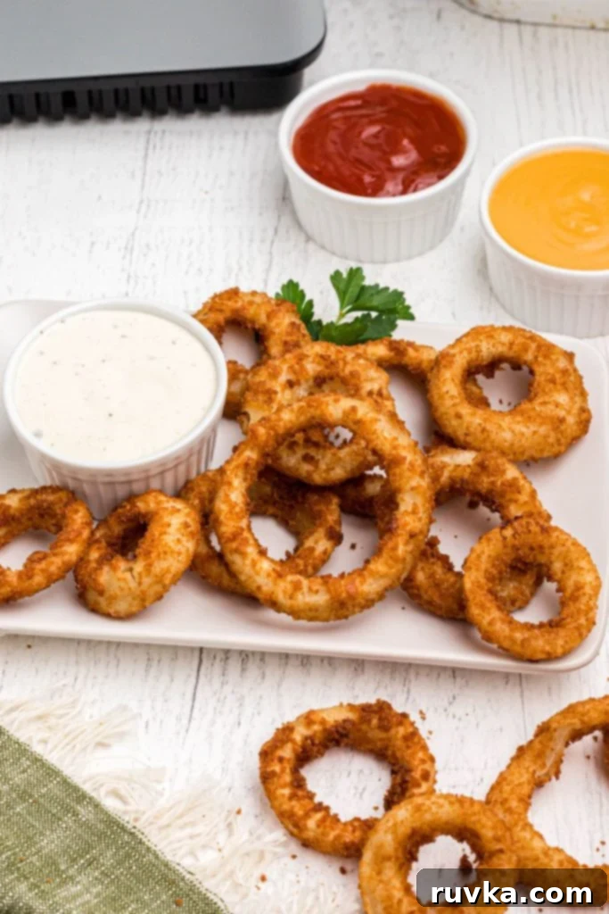 Effortlessly Crispy Air Fryer Onion Rings 6 Golden crispy air fried onion rings stacked on a white plate served with ranch and other sauces.