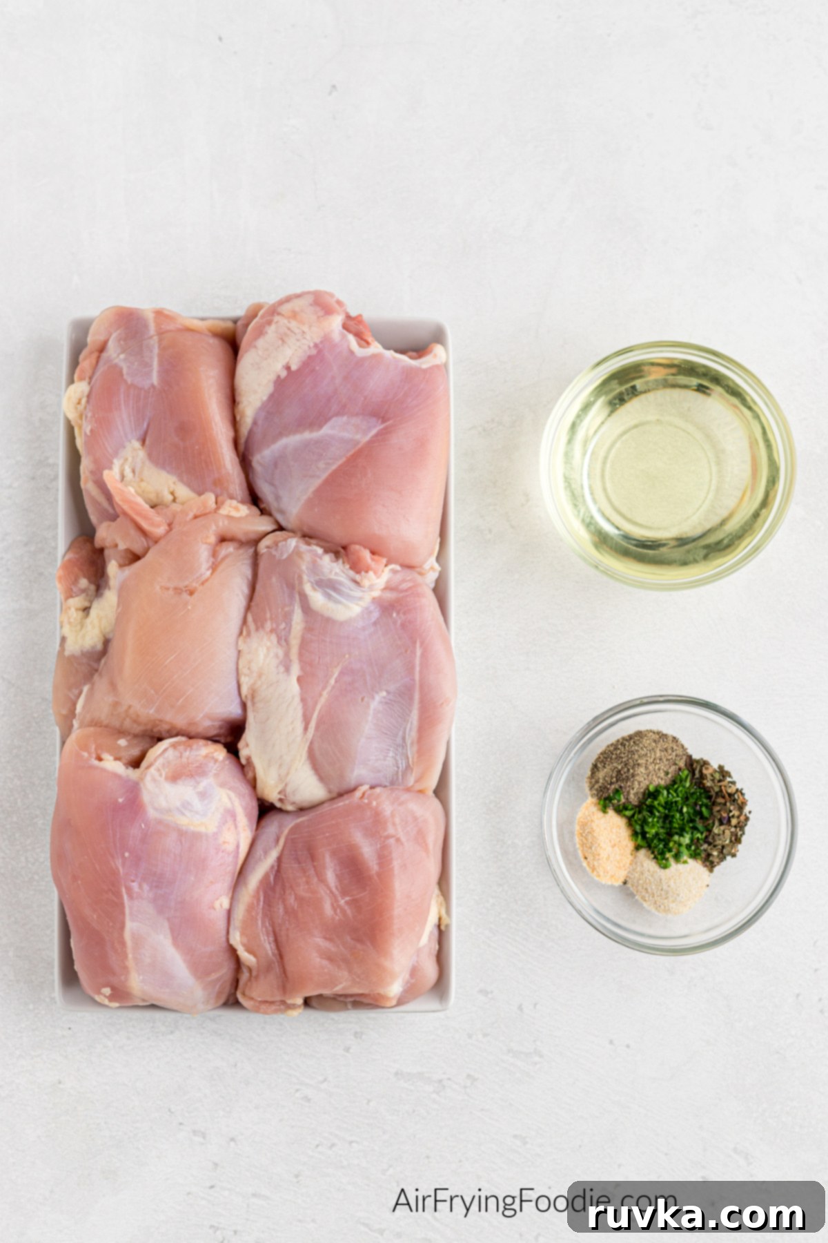Crispy Air-Fried Chicken Thighs 3 raw boneless, skinless chicken thighs, a small bowl of oil, and a small bowl of seasonings all on a white board.