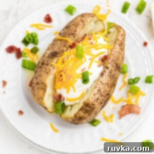 Crispy Air Fryer Loaded Potatoes 8 Baked potato stuffed with cheese, sour cream, bacon crumbles, on a white plate with chopped green onion and crumbles scattered on the table.