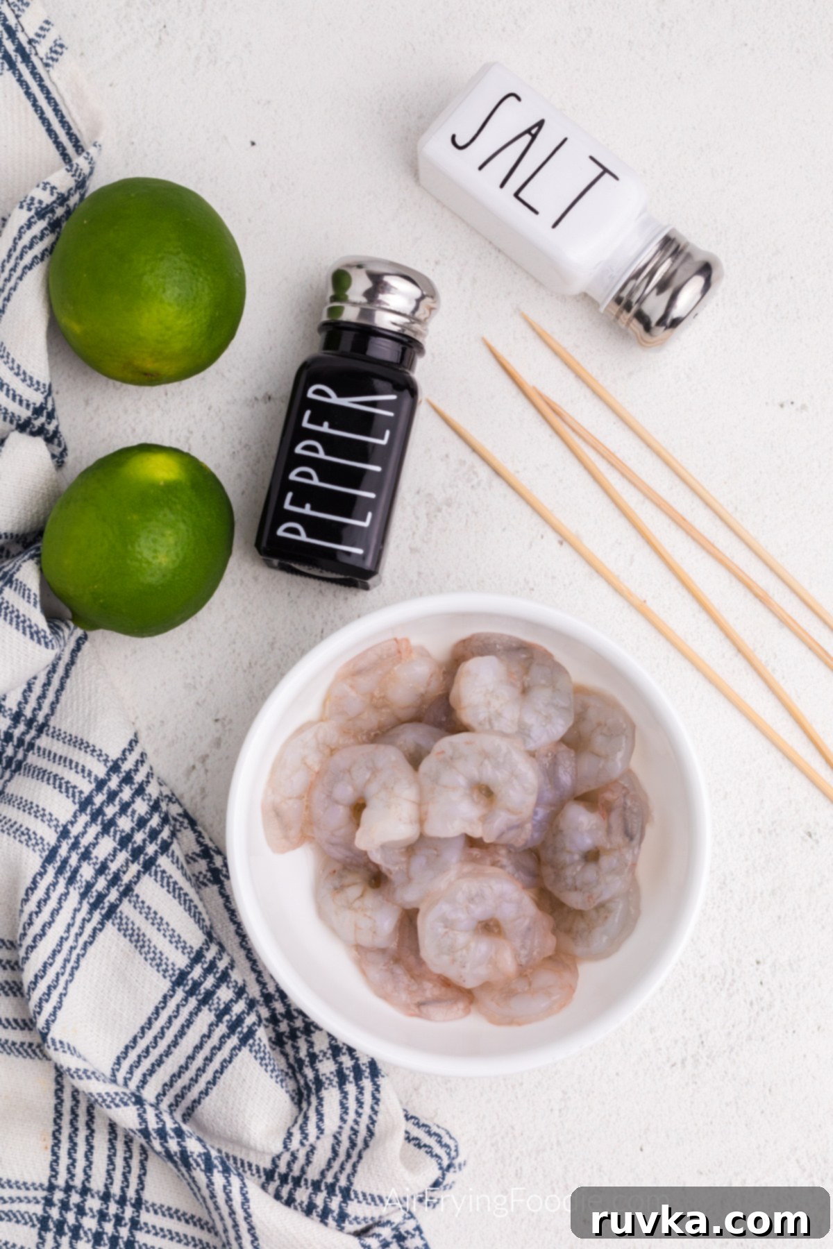 Easy Air Fryer Shrimp Skewers 3 Shrimp, limes, wooden skewers, and salt and pepper on a table.