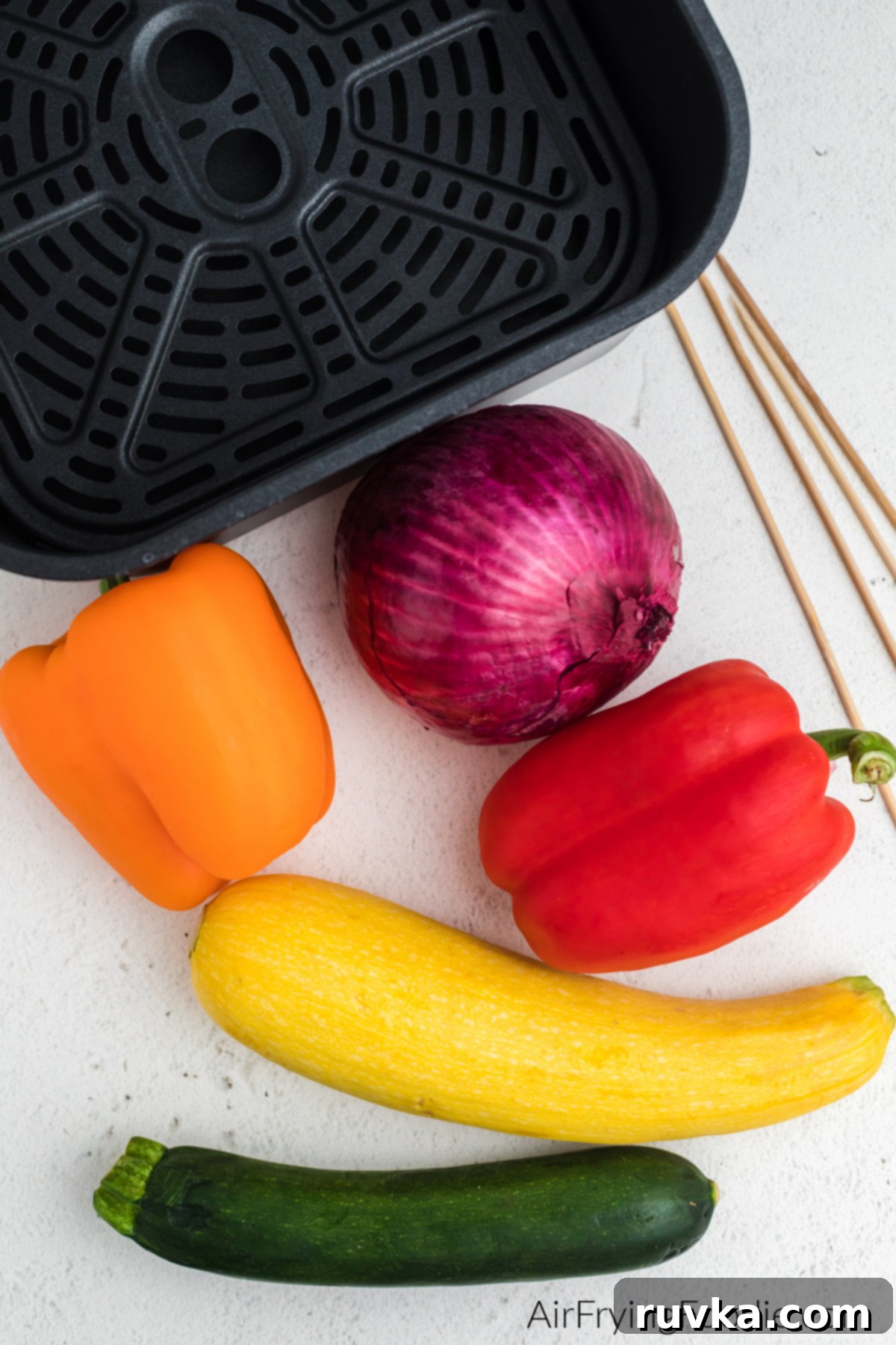 Crispy Air Fryer Vegetable Skewers 3 Freshly cut orange and red bell pepper, red onion, yellow squash, and zucchini squash arranged with wooden skewers and an air fryer basket, ready for assembly.