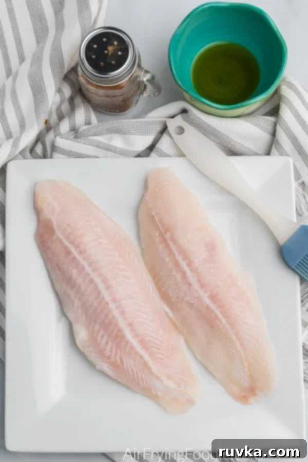 Air Fryer Swai Fish 4 uncooked swai fillets in a white plate with a brush, and blackened seasoning and olive oil above.