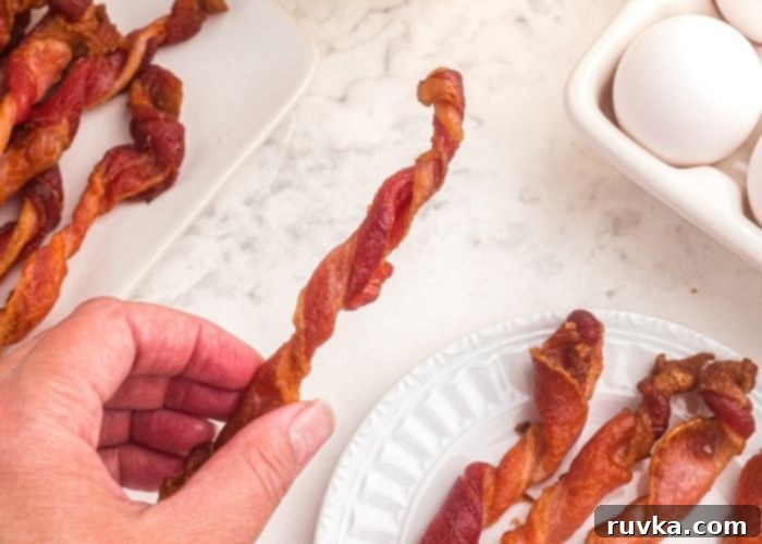 Air Fryer Twisted Bacon 7 A hand holding a piece of twisted bacon over a plate of bacon on a white marble table.