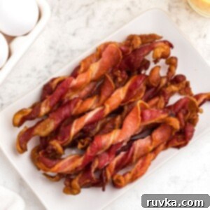 Air Fryer Twisted Bacon 8 Crispy twisted bacon, stacked and served on a white square plate, next to orange juice and eggs.