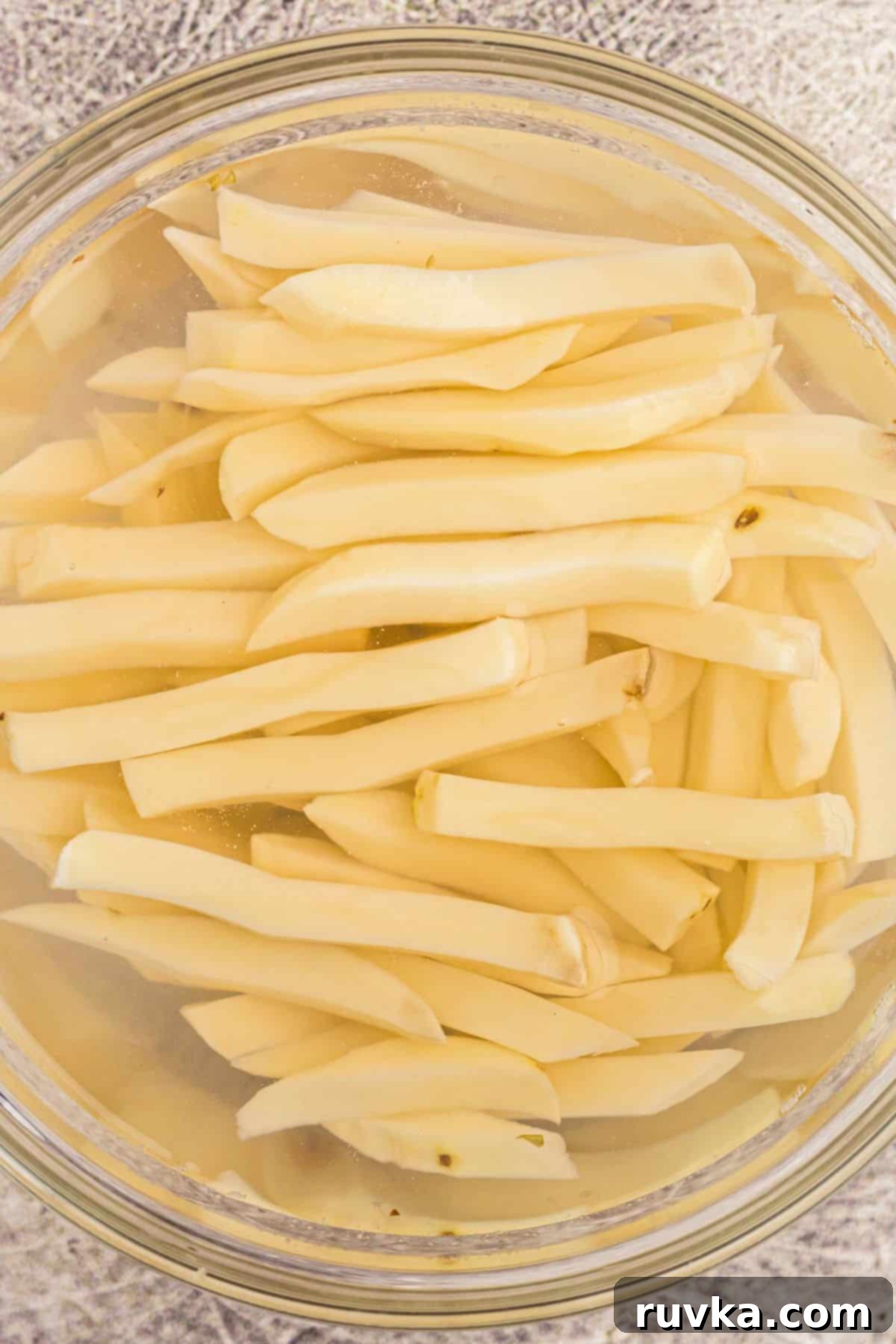 Crispy Air Fryer French Fries 5 Potato slices cut into fries, being soaked in a clear glass bowl filled with cold water.
