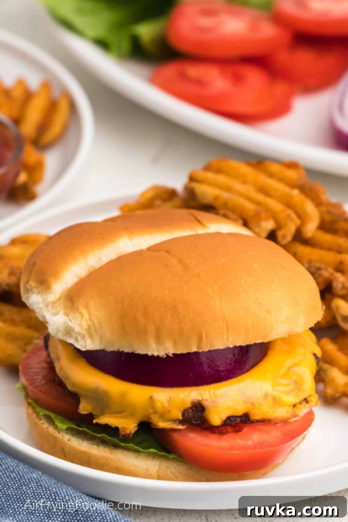 Perfectly Reheat Your Burger in the Air Fryer 4 reheated burger in air fryer that has been topped with cheese, onion, and served on a bun with lettuce and tomato and on a plate with a side of waffle fries.