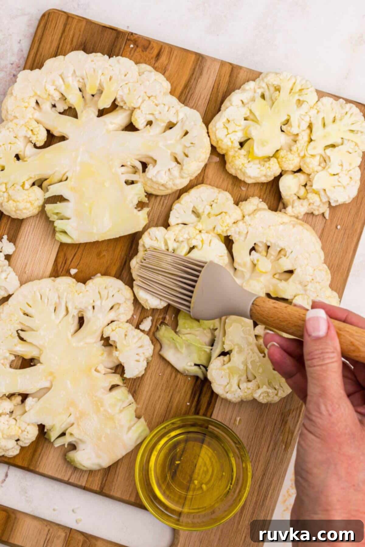 Crispy Air Fryer Cauliflower Steaks 5 Uncooked cauliflower steaks on a wooden cutting board being brushed with olive oil.