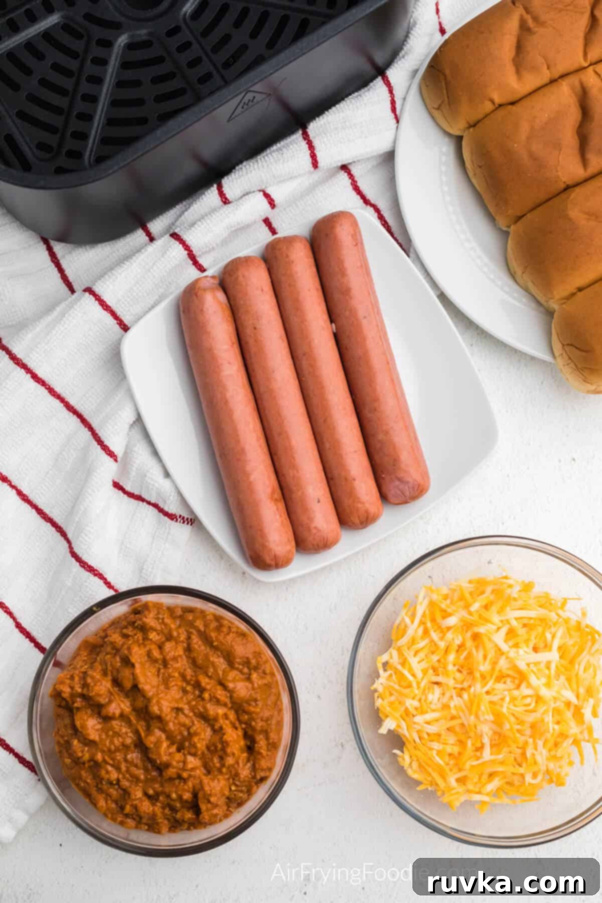 Crispy Air Fryer Chili Cheese Dogs 3 Ingredients needed to make chili cheese dogs in the air fryer on a table. Hotdogs on a plate, buns on a plate, chili in a bowl, and shredded cheese in a bowl, neatly arranged.
