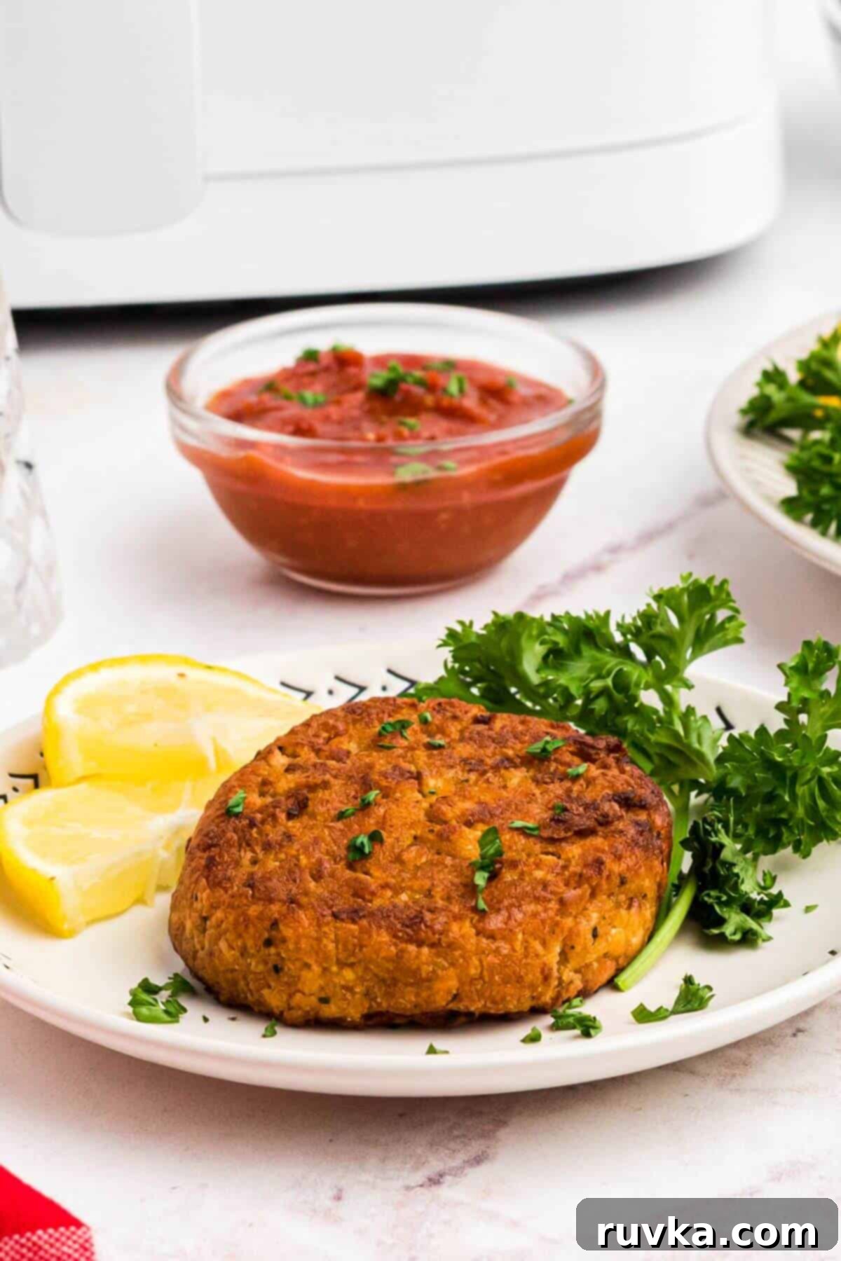Crispy Air Fryer Frozen Crab Cakes 2 golden brown fried crab cakes on a small plate with parsley and lemon in front of an air fryer.