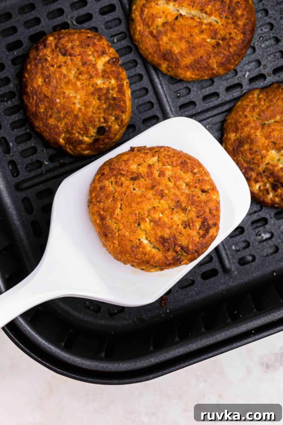 Crispy Air Fryer Frozen Crab Cakes 5 Golden crispy crab cakes in the air fryer basket with one being lifted by a white spatula.