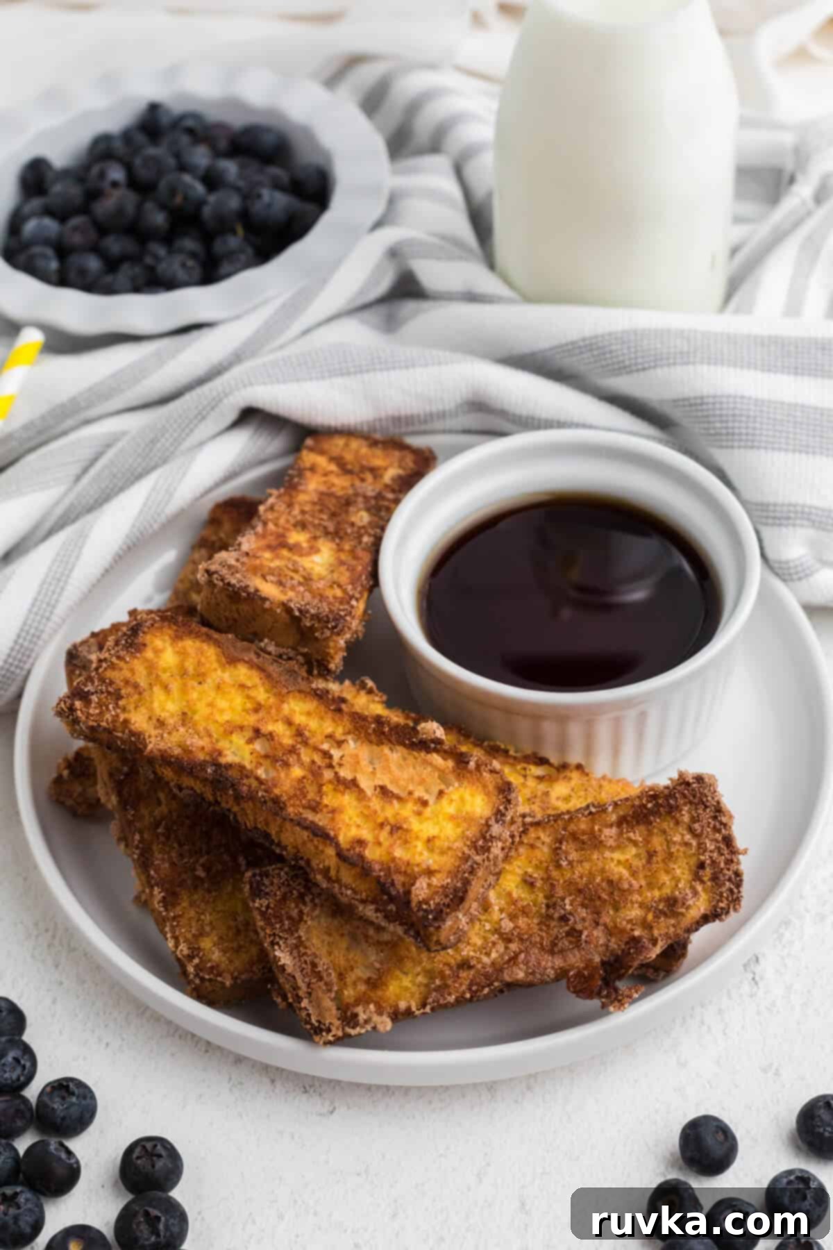 Golden Air Fryer French Toast Sticks 2 French toast sticks made in the air fryer, served on a white plate with a bowl of syrup for dipping.