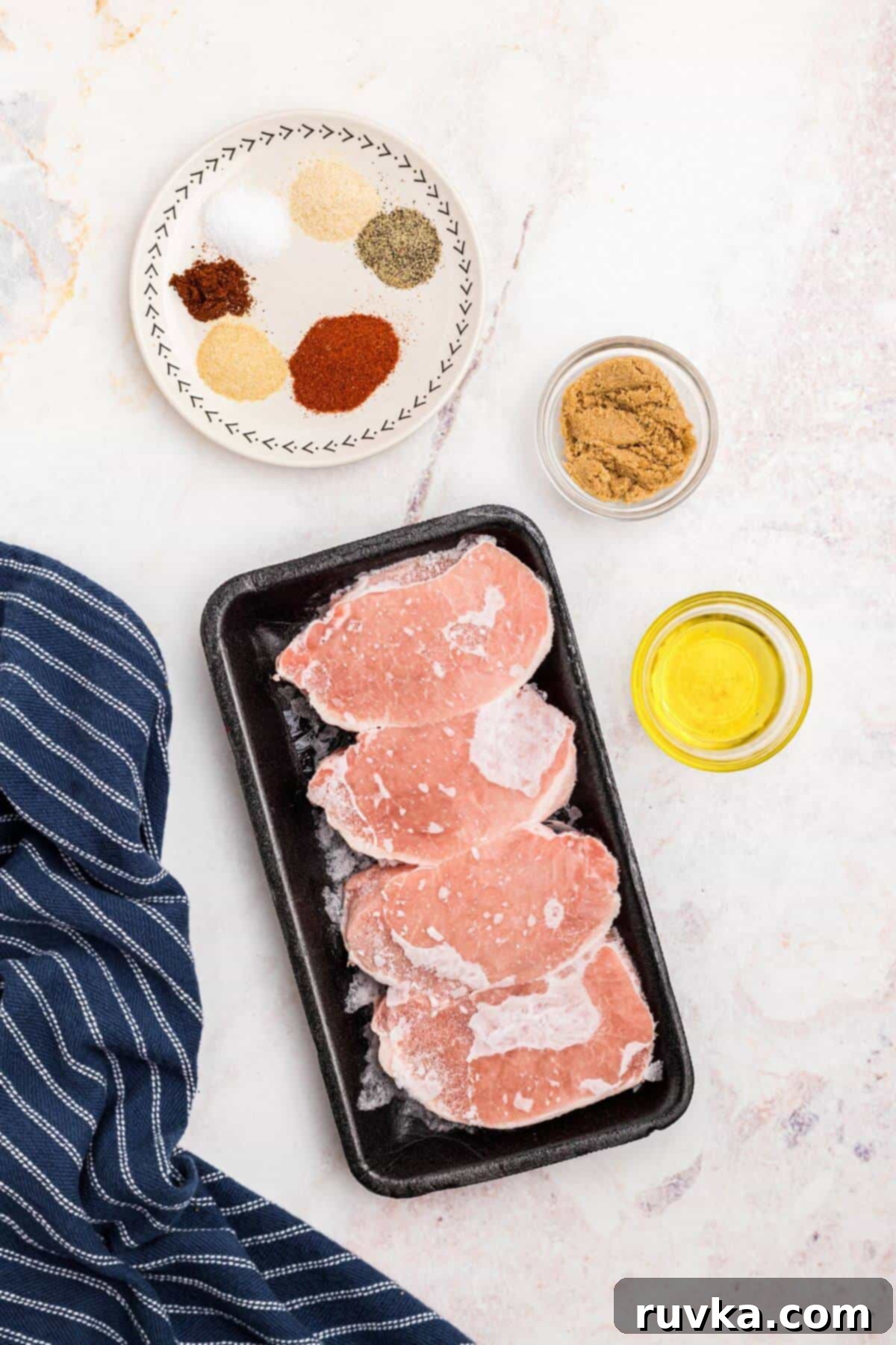 No-Thaw Air Fryer Pork Chops 3 Frozen pork chops on a marble table with olive oil and seasonings for the rub measured out in small dishes.
