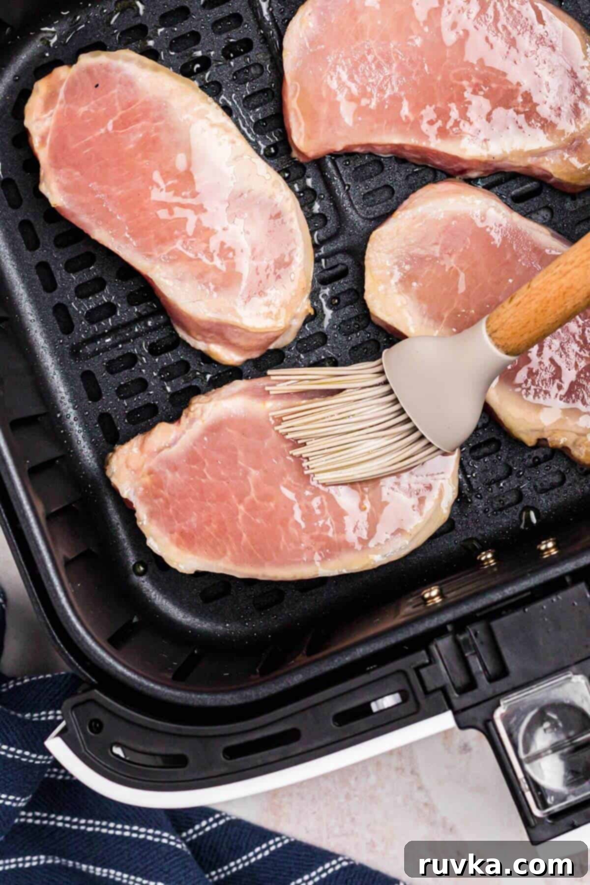 No-Thaw Air Fryer Pork Chops 6 Uncooked pork chops in the air fryer basket being brushed with olive oil before being seasoned.