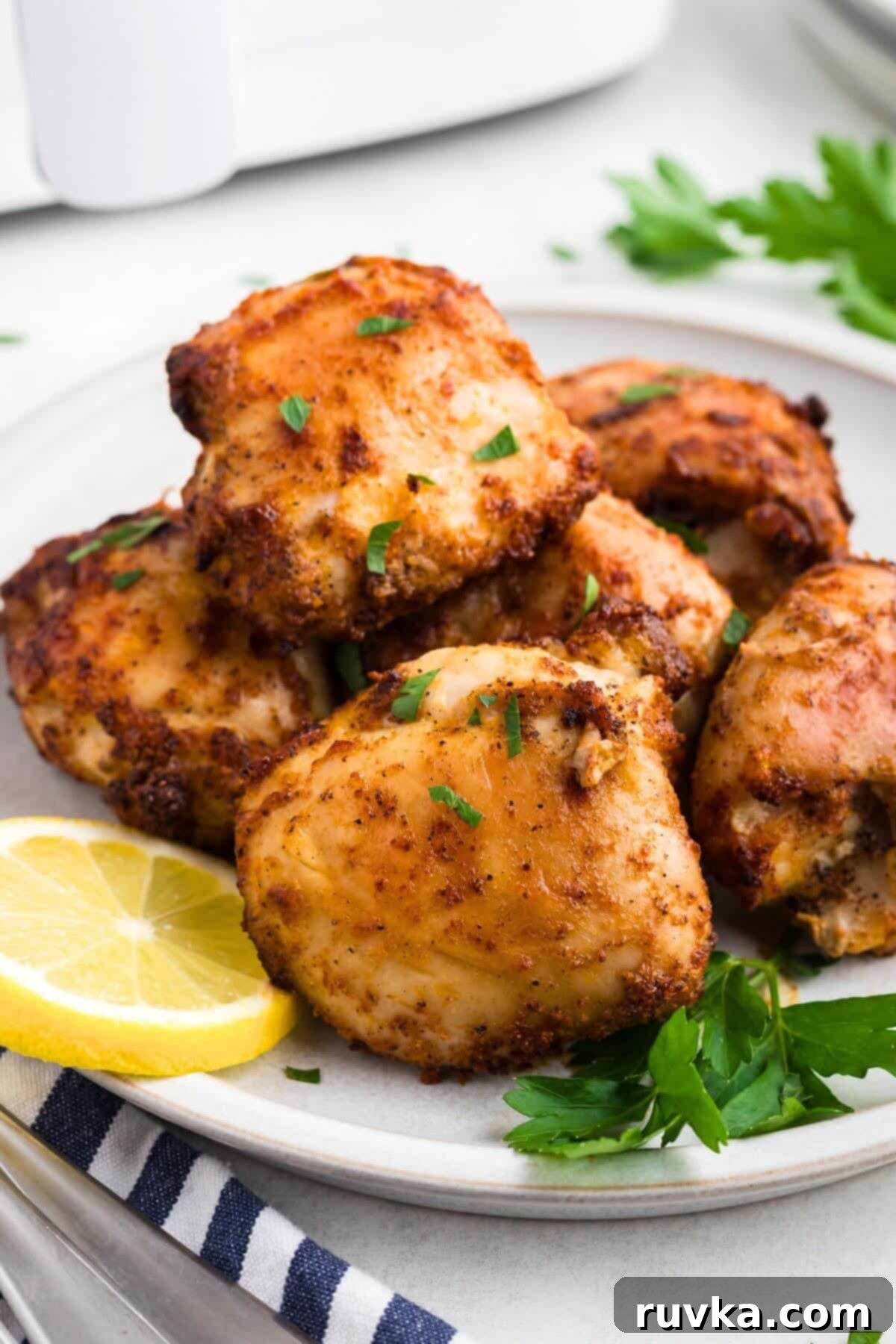 Freezer to Fabulous Air Fryer Chicken Thighs 2 Crispy brown seasoned chicken thighs on a round white plate with lemon slices and parsley garnish.