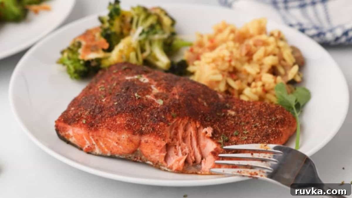 15 Foolproof Air Fryer Recipes 6 Simple Salmon Filet seasoned and sir fried, then served on a white plate with a side of brocolli and rice in the background, with a fork at the front of the plate.