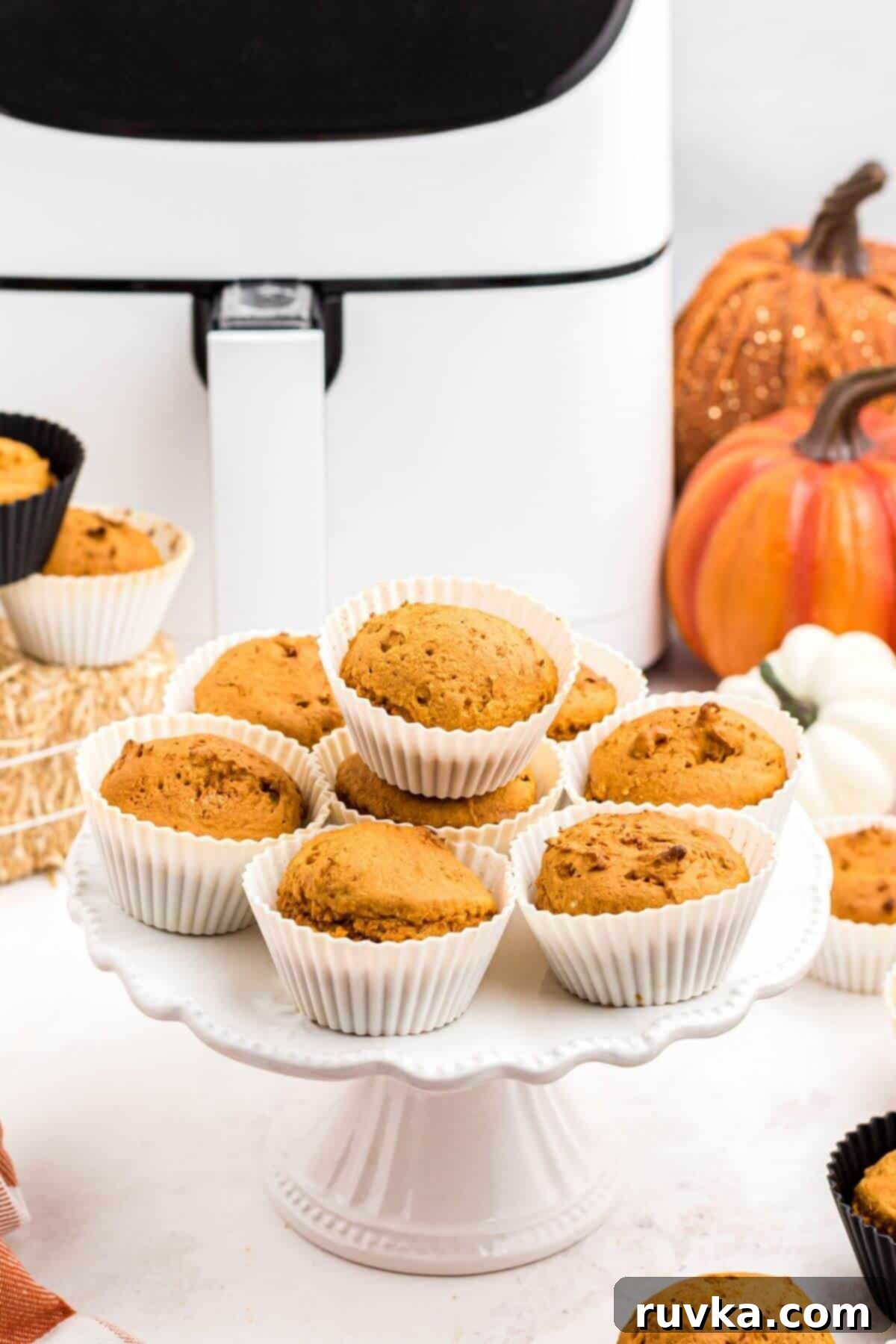 Easy Air Fryer 2 Ingredient Pumpkin Muffins 2 Golden orange pumpkin muffins on a cake stand, in front of the air fryer.