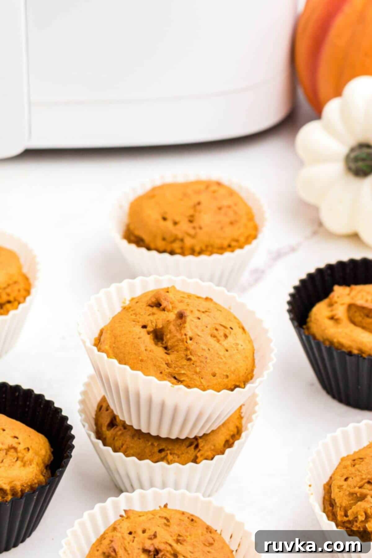 Easy Air Fryer 2 Ingredient Pumpkin Muffins 10 Pumpkin muffins scattered on a marble table in front of an air fryer, while still in their black or white silicone liners.