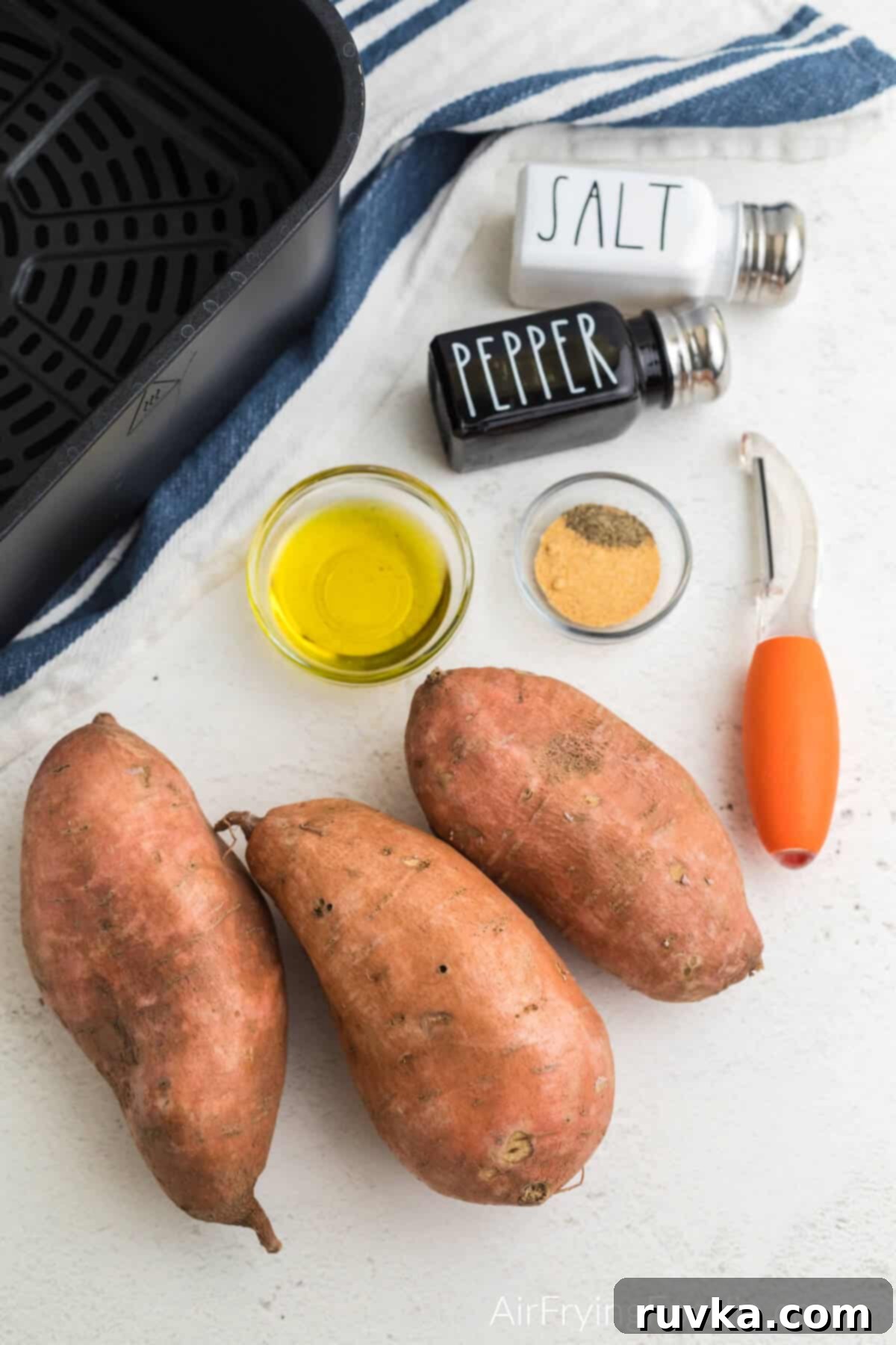 Crispy Air-Fried Sweet Potato Cubes 3 sweet potatoes, olive oil, garlic powder, pepper, an air fryer basket, and vegetable peeler on a white table.