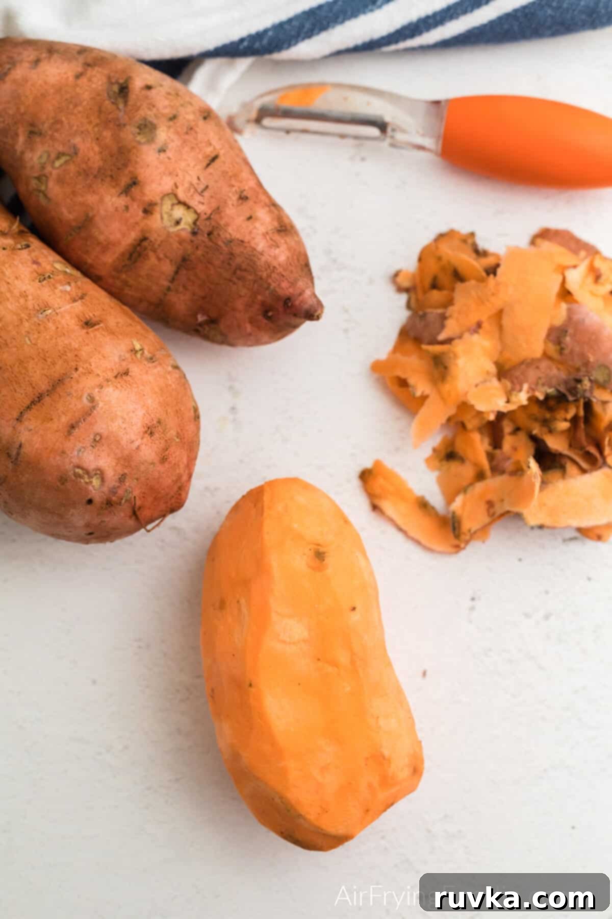 Crispy Air-Fried Sweet Potato Cubes 4 Peeled sweet potato on a table top surrounded by sweet potatoes and peelings with a vegetable peeler off to the side.