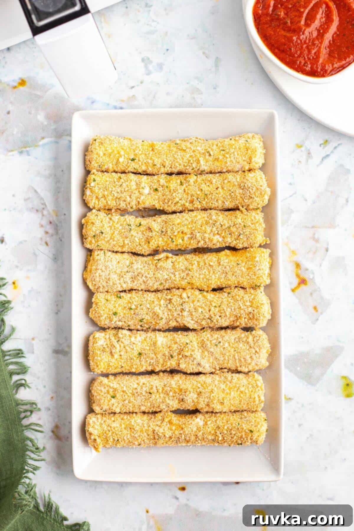 Crispy Air Fryer Mozzarella Sticks 5 Mozzarella cheese sticks coated in a breaded coating on a white rectangle plate before being air fried.