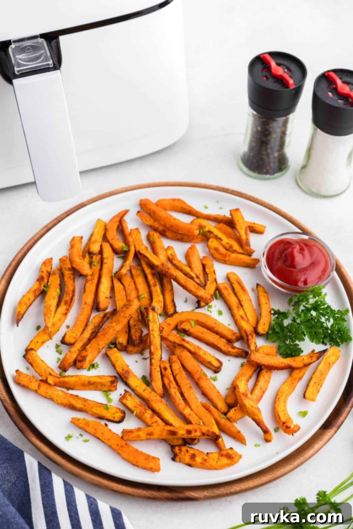 Crispy Air Fryer Sweet Potato Fries 2 Orange golden sweet potato fries on a round white plate with a small dish of ketchup in front of a white air fryer.