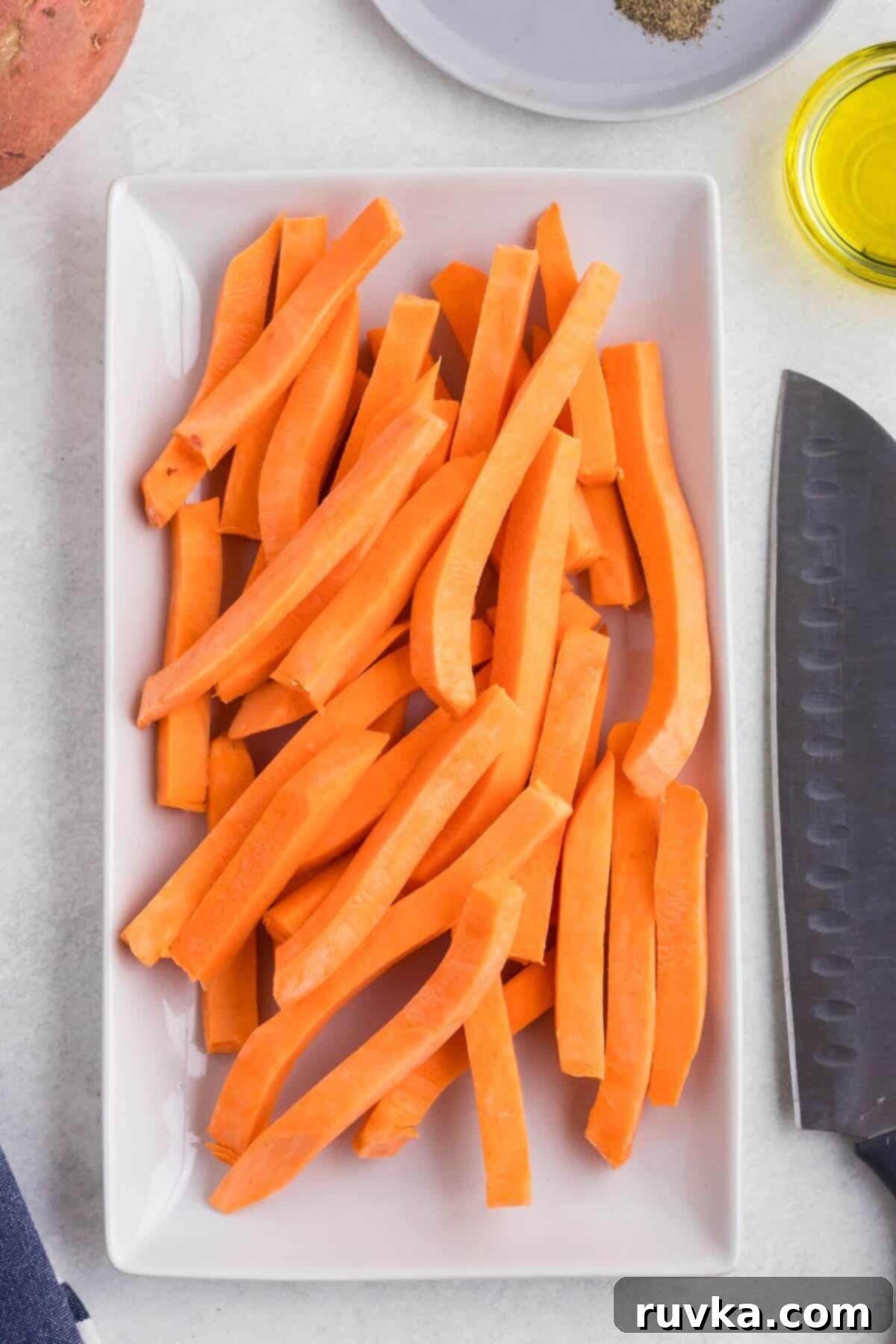 Crispy Air Fryer Sweet Potato Fries 4 Sweet potatoes cut into slices on a white rectangle plate with olive oil and seasonings on a white table.