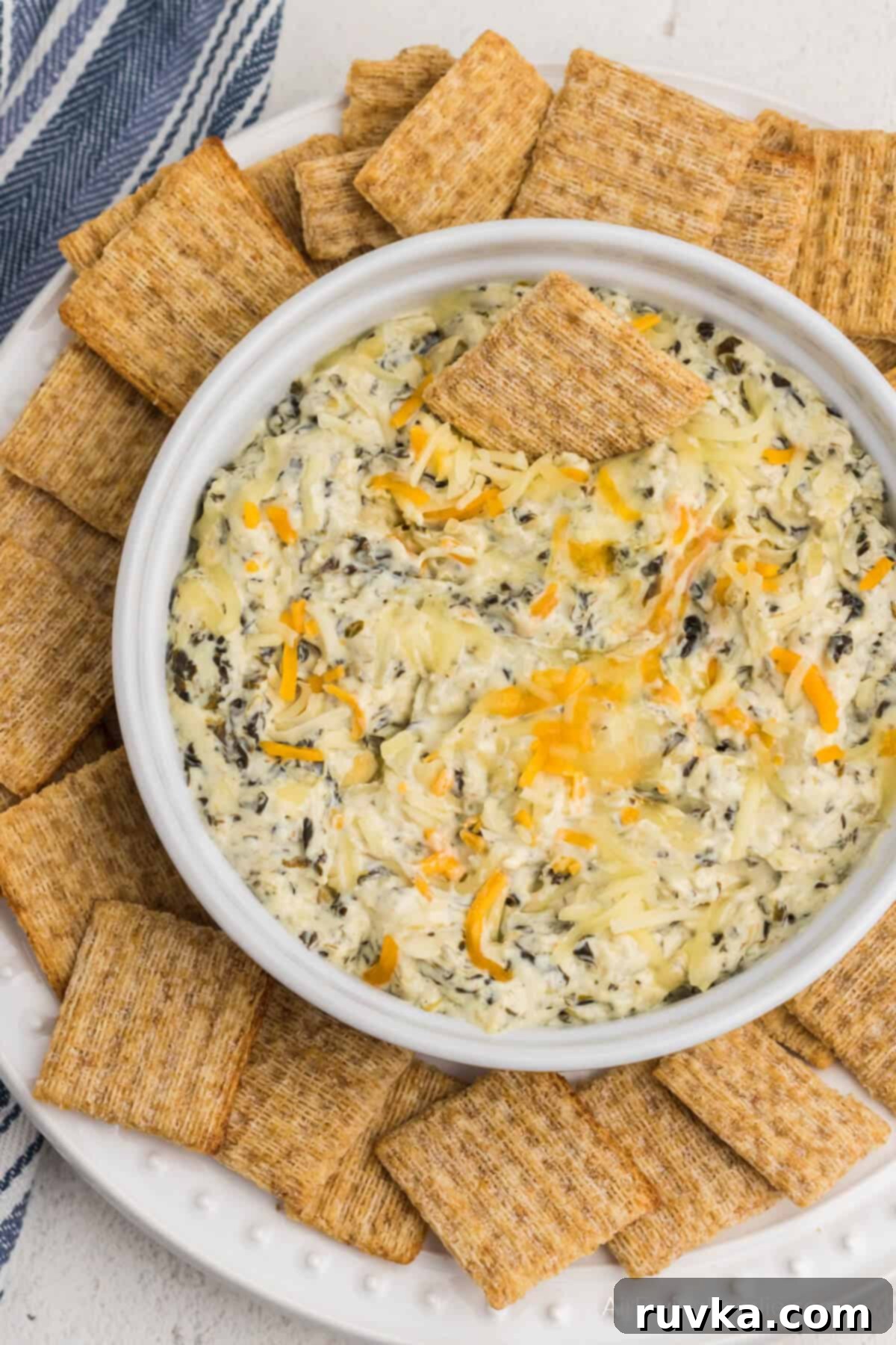 Easy Air Fryer Artichoke Spinach Dip 4 Air fryer artichoke dip topped with cheese and in an air fryer safe dish, surrounded by Triscuit crackers for dipping.